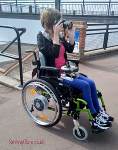 Slim woman in a green framed wheelchair, wearing bright blue trousers, a pink top and black hooded jacket, holding a camera up to her face taking a photograph